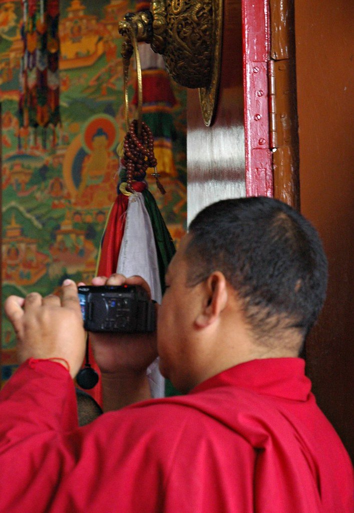 What a Tibetan Monk Taught Me About the Purpose of Technology - Monk with camera getting in a few shots, his mala tied in a double dorje above him, Buddha painted on the far wall, mural, door ring, Tharlam Monastery, Boudha, Kathmandu, Nepal